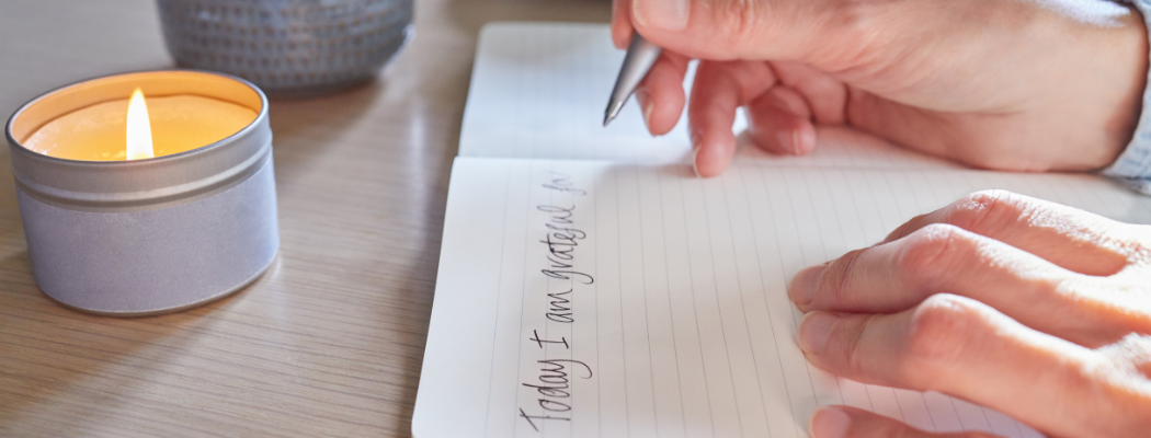 The hands of a young woman writing on a journal with a fountain pen. There is a small candle in a tin container by the journal.